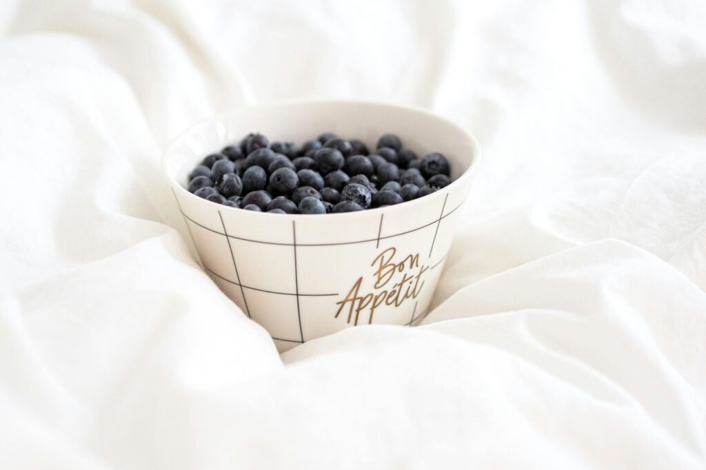 Picture of a white ceramic bowl with "Bon Appétit" written on it, filled with fresh blueberries resting on white bed linens.