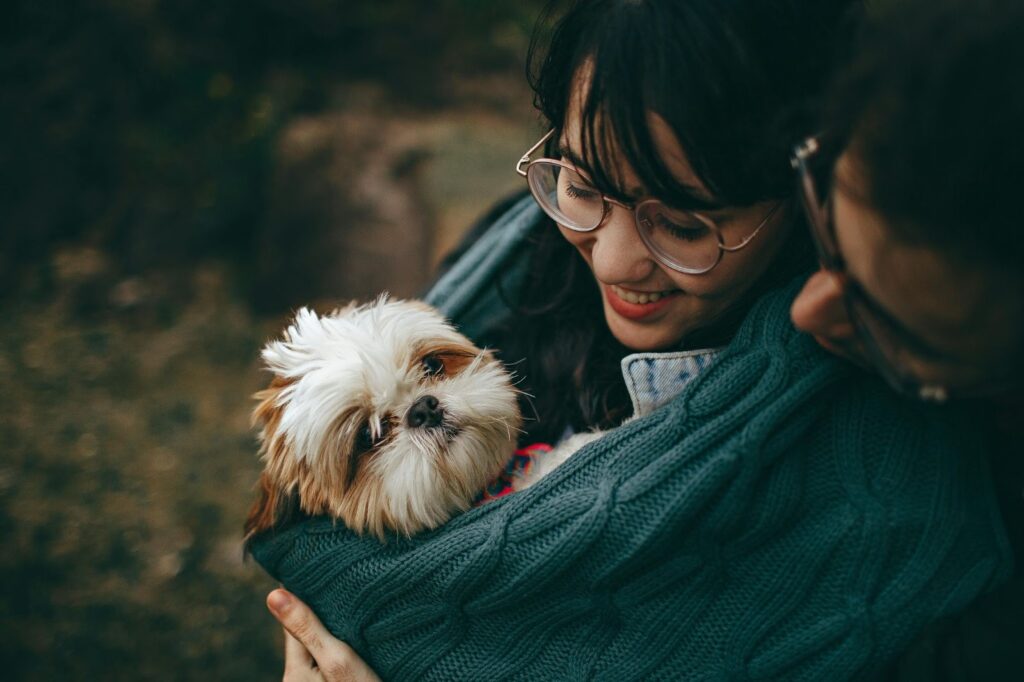 Picture of a woman smiling and holding a small shih tzu wrapped closely in a teal knitted blanket.