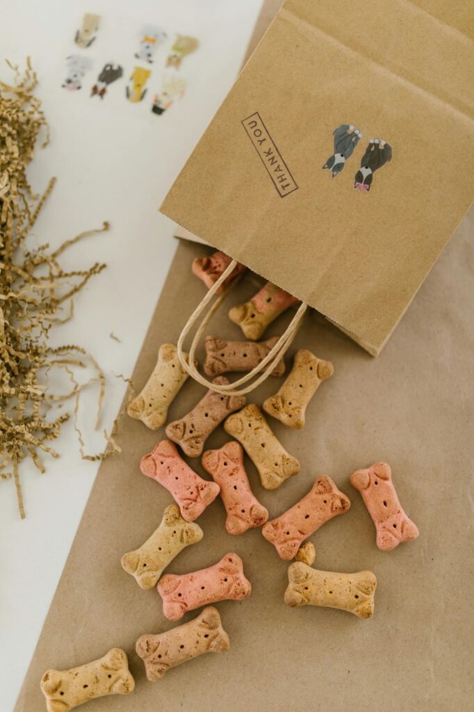 Picture of a brown paper gift bag overturned with bone-shaped dog treats in shades of tan and pink spilling out