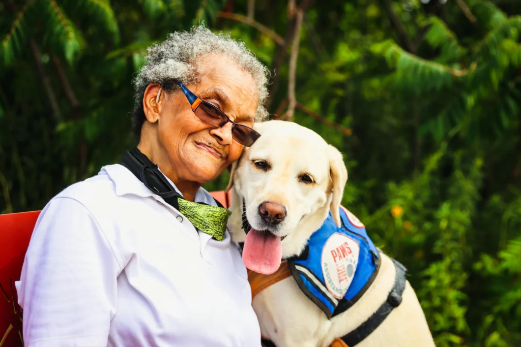Picture of a smiling woman embracing a yellow Labrador Retriever wearing a blue "PAWS" assistance vest