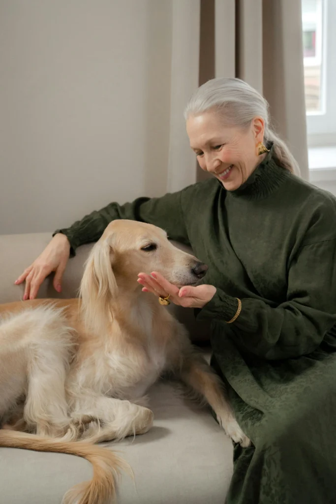 Picture of a woman in a green dress sitting on a sofa and gently offering her hand to a long-haired, light-coloured dog.