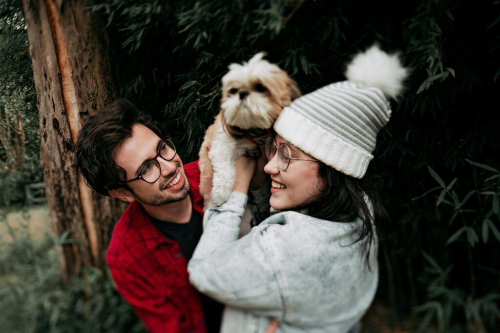 Picture of a smiling couple hugging a small fluffy brown and white dog outdoors.