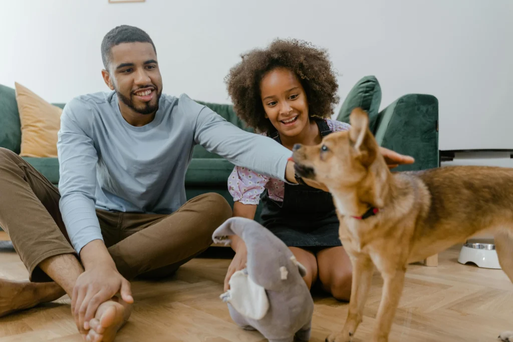 picture of a man and young girl sitting on a living room floor reaching out to pet a small brown dog