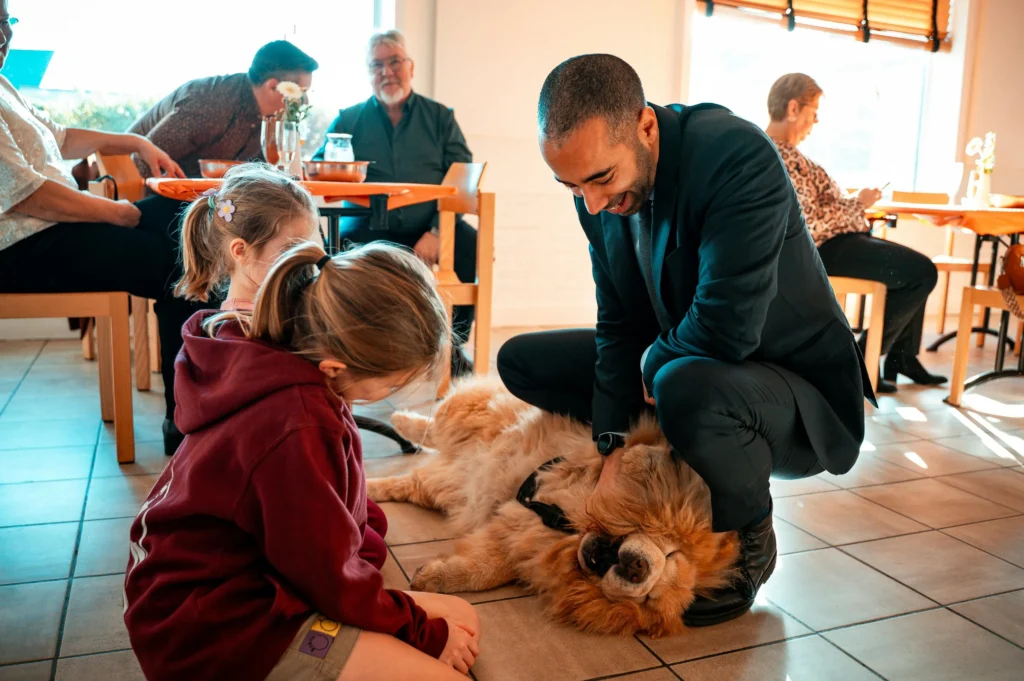 picture of a man in a suit kneeling down to rub the belly of a large golden fluffy dog near two children