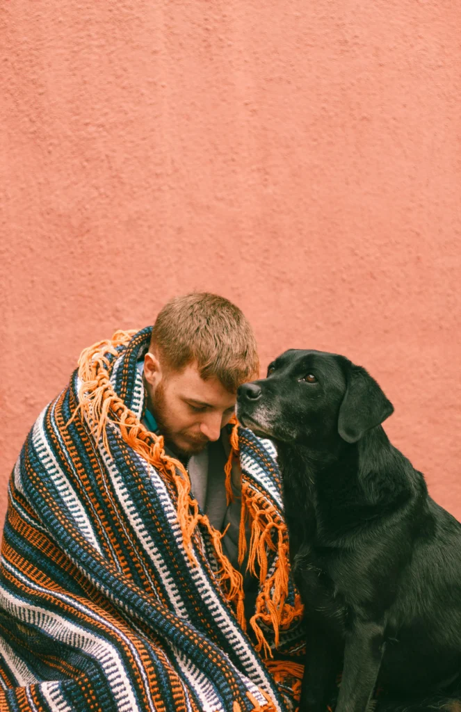 picture of a man wrapped in a colorful patterned blanket sitting closely next to a black labrador retriever