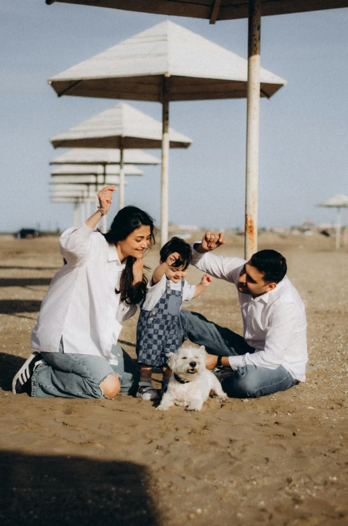 picture of a mother and father playing in the sand at the beach with their toddler and a small white terrier