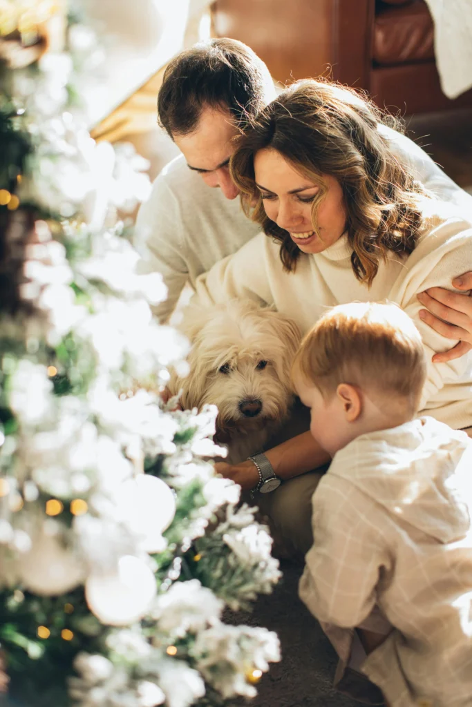 picture of a mother and father with their young son petting a fluffy white dog next to a decorated christmas tree