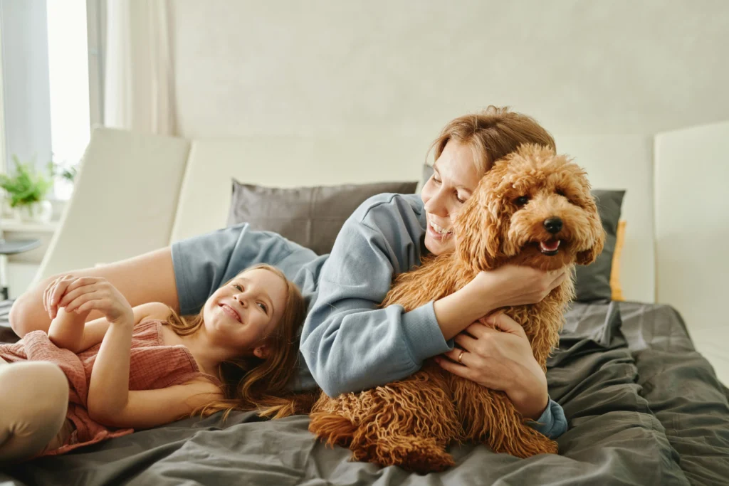picture of a woman and young girl laughing while cuddling a small brown fluffy poodle mix on a bed