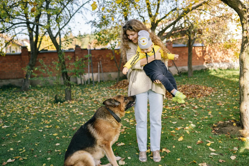 Picture of a woman holding a young child while a German Shepherd looks up at them in an autumn yard