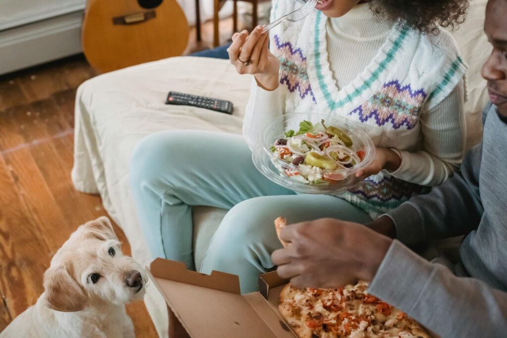 Picture of a small white dog watching two people eat pizza and salad.
