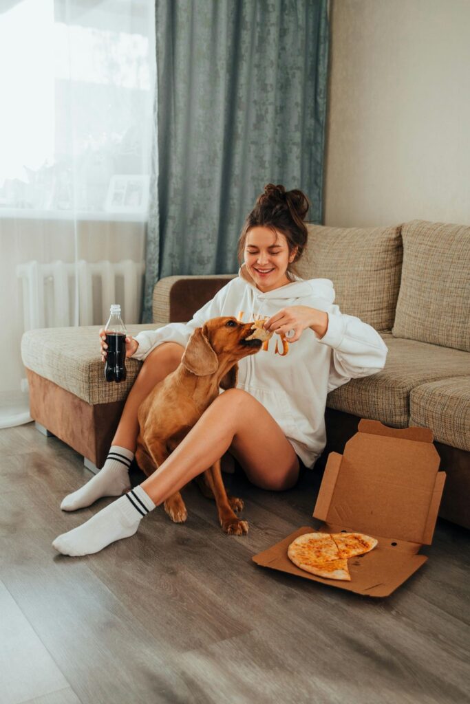 Picture of a happy woman sitting by a couch while her dog takes a bite of pizza.
