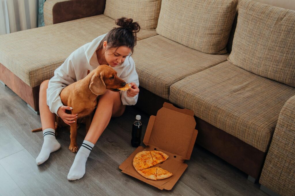 Picture of a woman sitting on the floor feeding a slice of pizza to a brown dog.