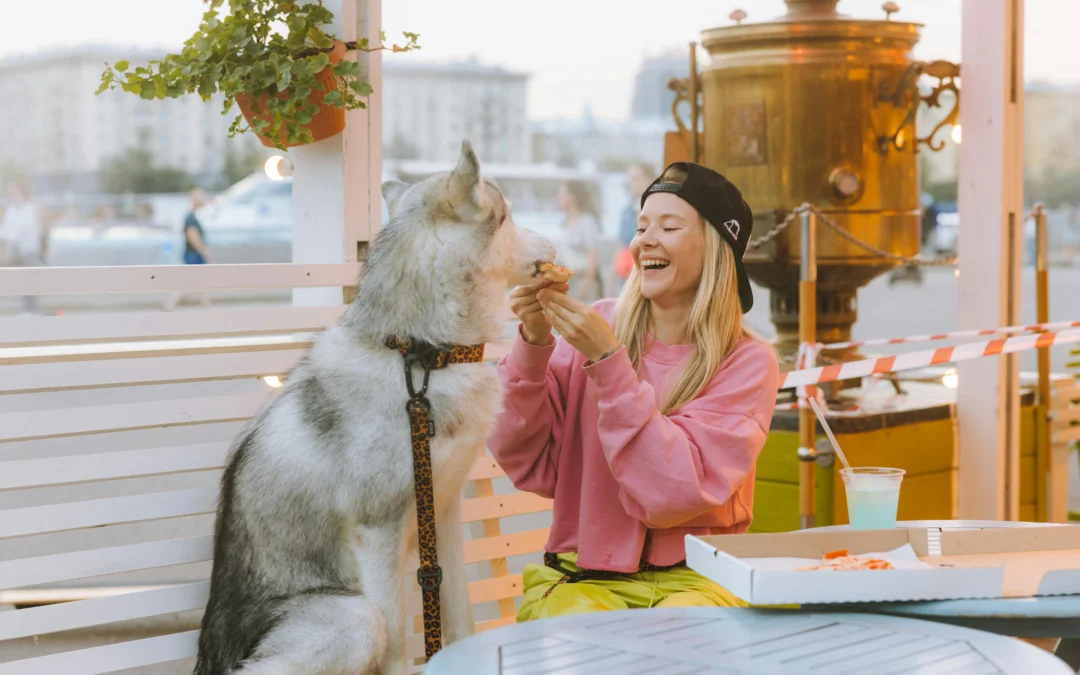 Picture of a smiling woman feeding a piece of pizza to a husky at an outdoor patio