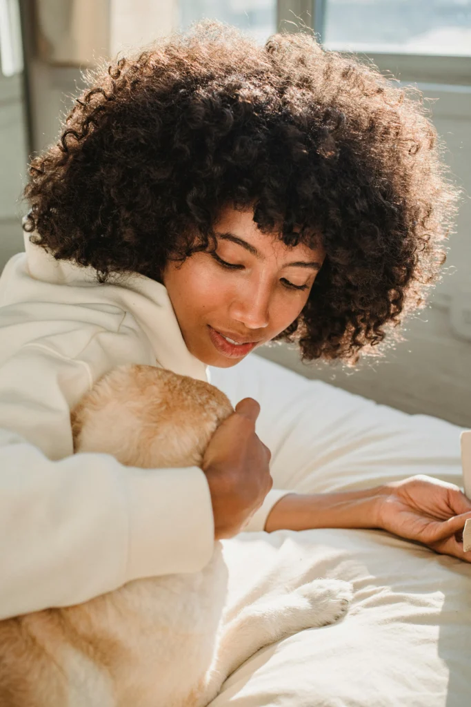 picture of a woman with curly hair cuddling a light colored dog while lying on a bed in the sunlight