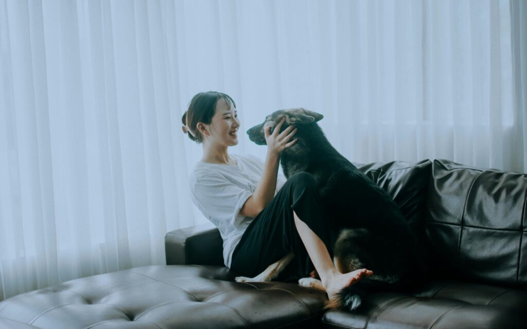 Picture of a smiling woman sitting near a leather couch, playing with a black dog.