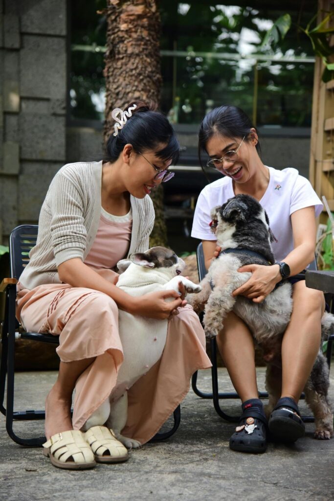 Picture of a couple of women sitting outdoors, smiling and petting two small dogs.