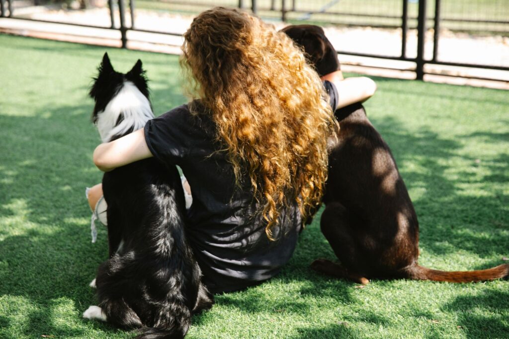 Picture of a woman seen from behind, hugging two dogs on the grass
